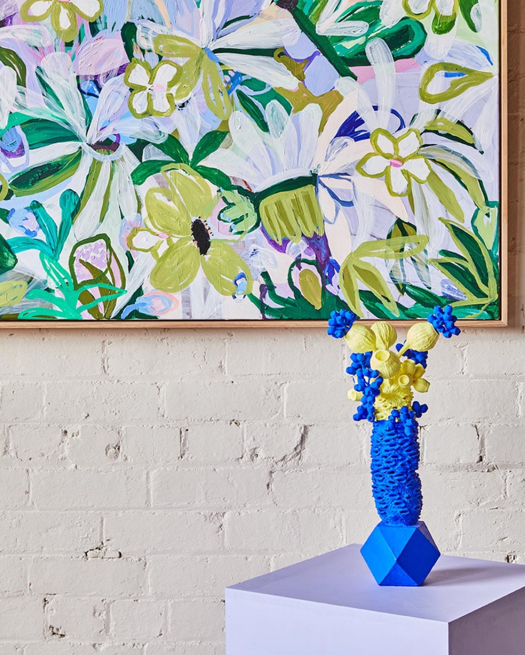 Blue and yellow vase on a white pedestal in front of a floral painting on a brick wall.